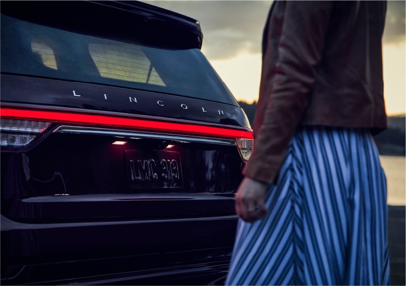 A person is shown near the rear of a 2023 Lincoln Aviator® SUV as the Lincoln Embrace illuminates the rear lights | J.C. Lewis Lincoln in Savannah GA