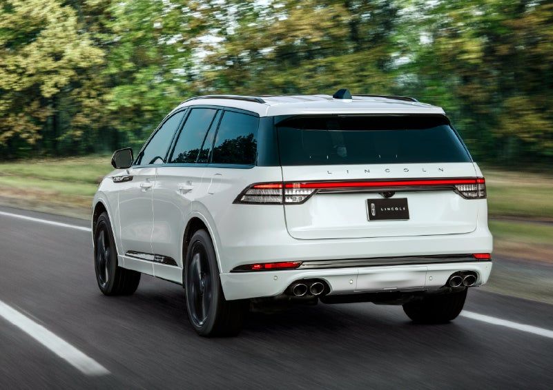 A white 2026 Lincoln Aviator® SUV is being driven away down a road lined with colorful trees. | J.C. Lewis Lincoln in Savannah GA